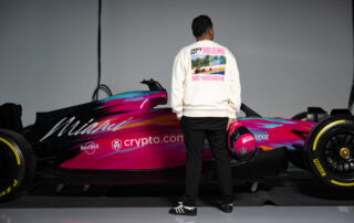 A model standing in front of a Miami pink f1 car displays the back of his sweatshirt. New merch available.