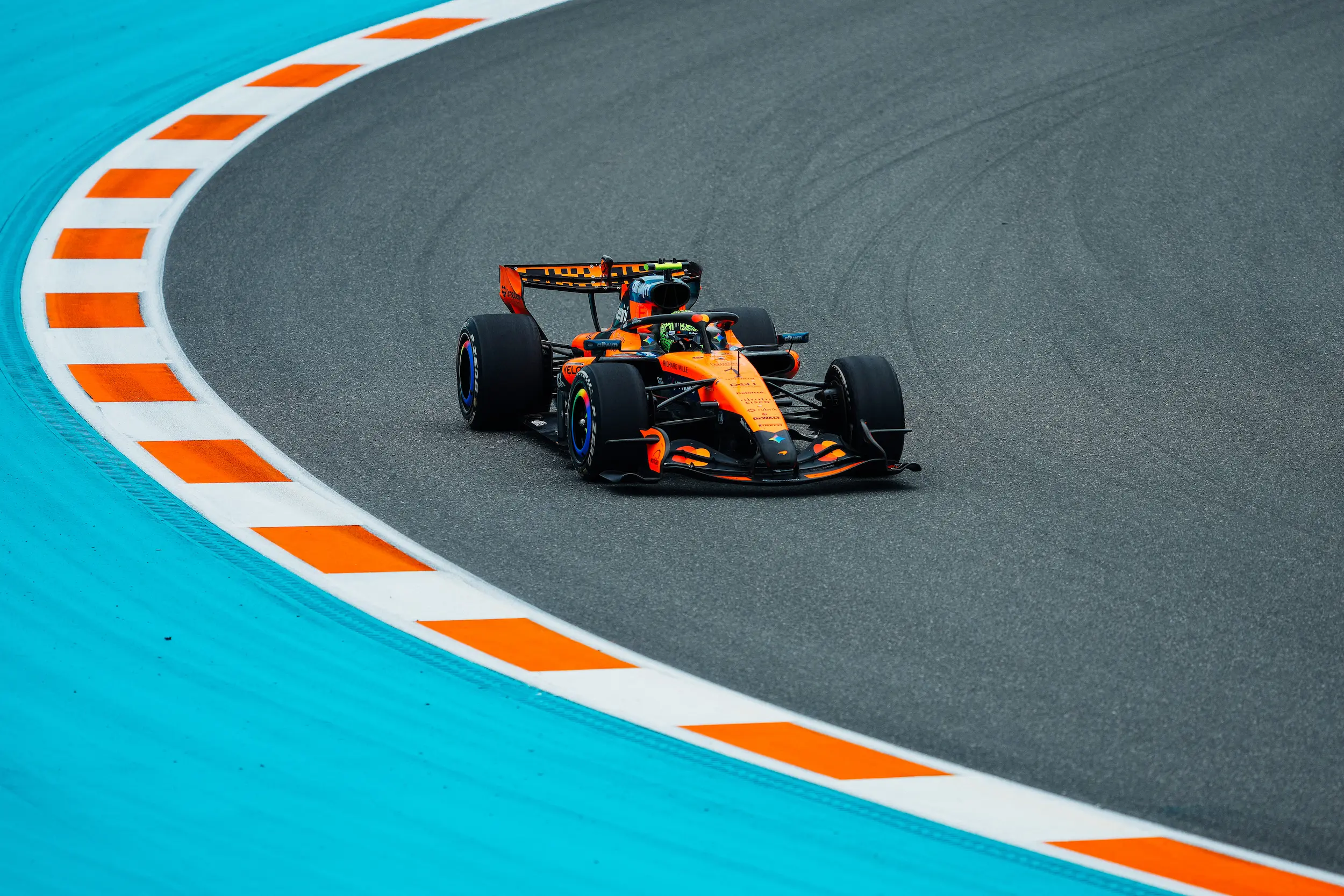 260503_F1MGP_Getty2274325819 (1) Orange Formula 1 race car navigating a sweeping turn on a racetrack, bordered by bright turquoise runoff areas and orange-and-white curbing. The car is positioned near the apex of the corner on a dry asphalt surface.