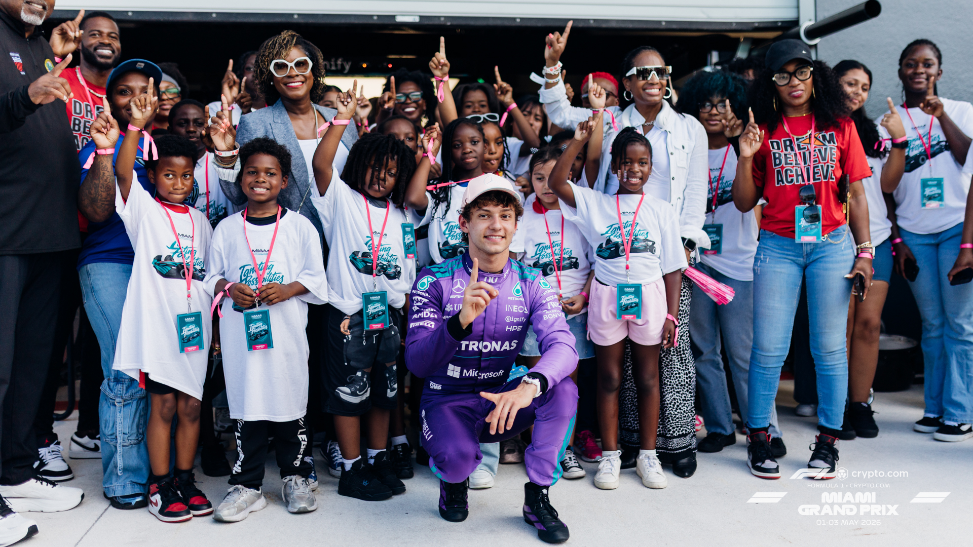 A large group of children and adults wearing white event T‑shirts and pink lanyards stand together with raised index fingers, while a driver in a purple racing suit kneels in front of the group; the setting appears to be a motorsport venue with signage reading “crypto.com Formula 1 Miami Grand Prix 01–03 May 2026.”
